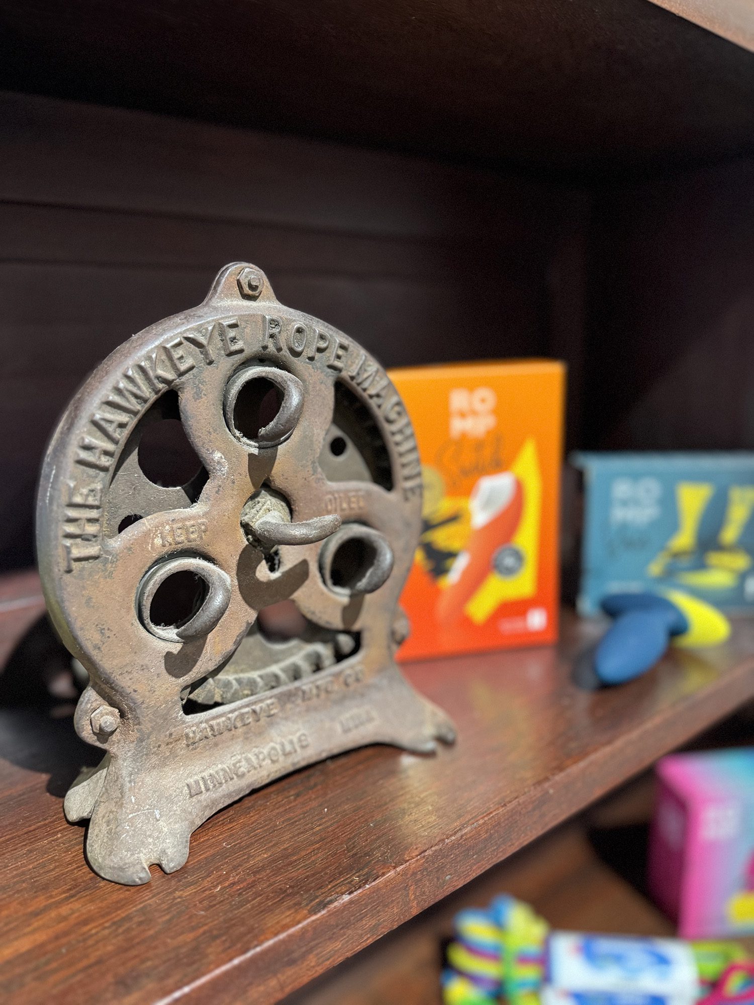 The Hawkeye Rope Mag-hinge, in the foreground of a close up photo of Studio Allegory Products sitting on a wooden shelf. Behind the mag-hinge, other colourful sex toys are slightly out of focus.